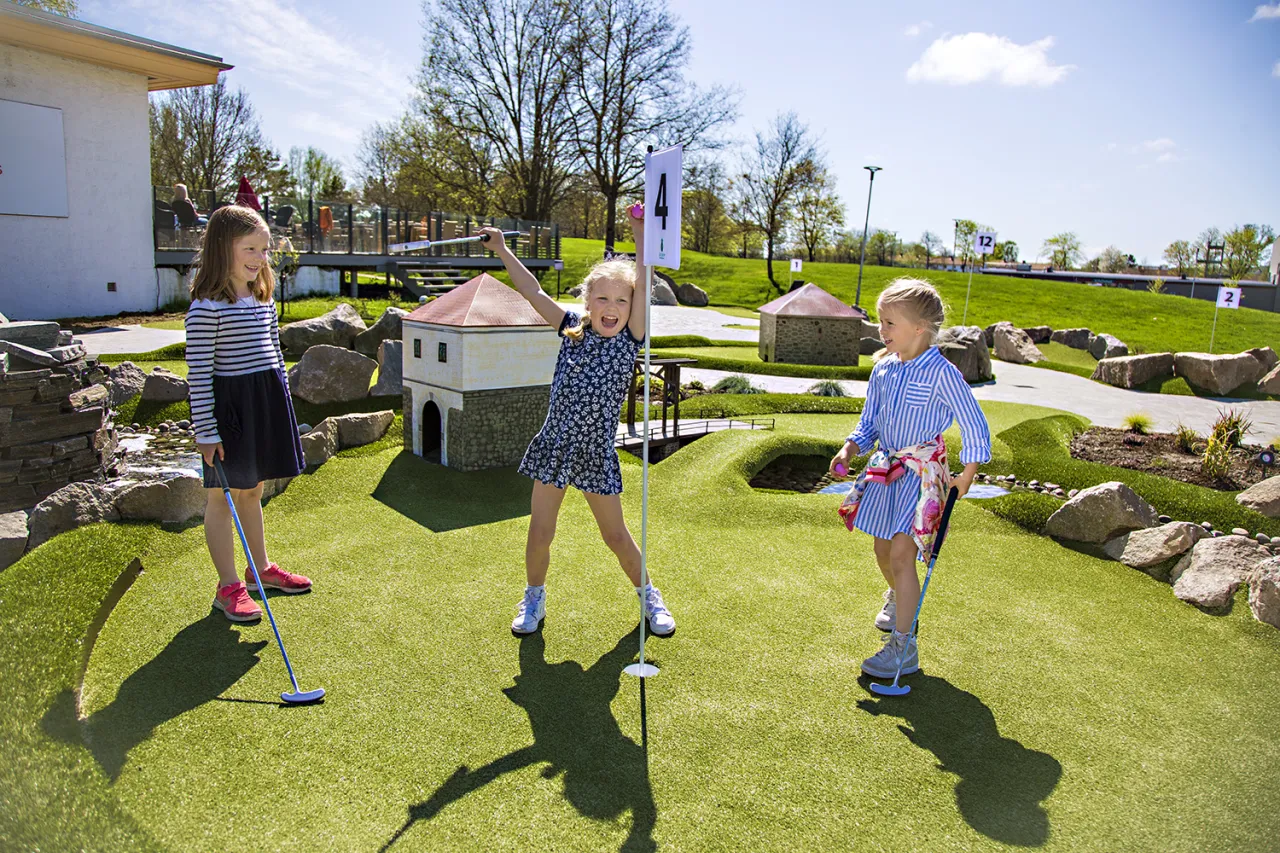 Tre jenter med golfkøller står på en minigolfbane ved flagg og hull, med steiner og små bygg i bakgrunnen.