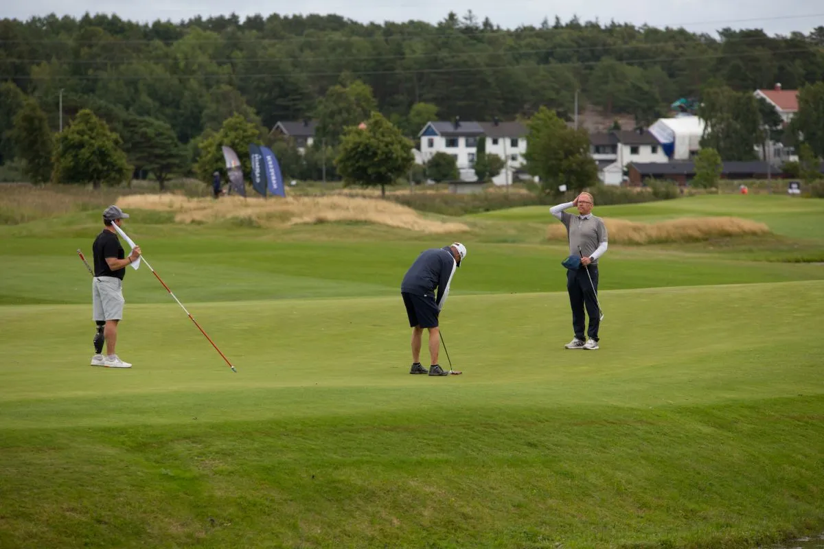 Tre golfspillere på en green; én putter mens to står og ser på, med trær og hus i bakgrunnen.