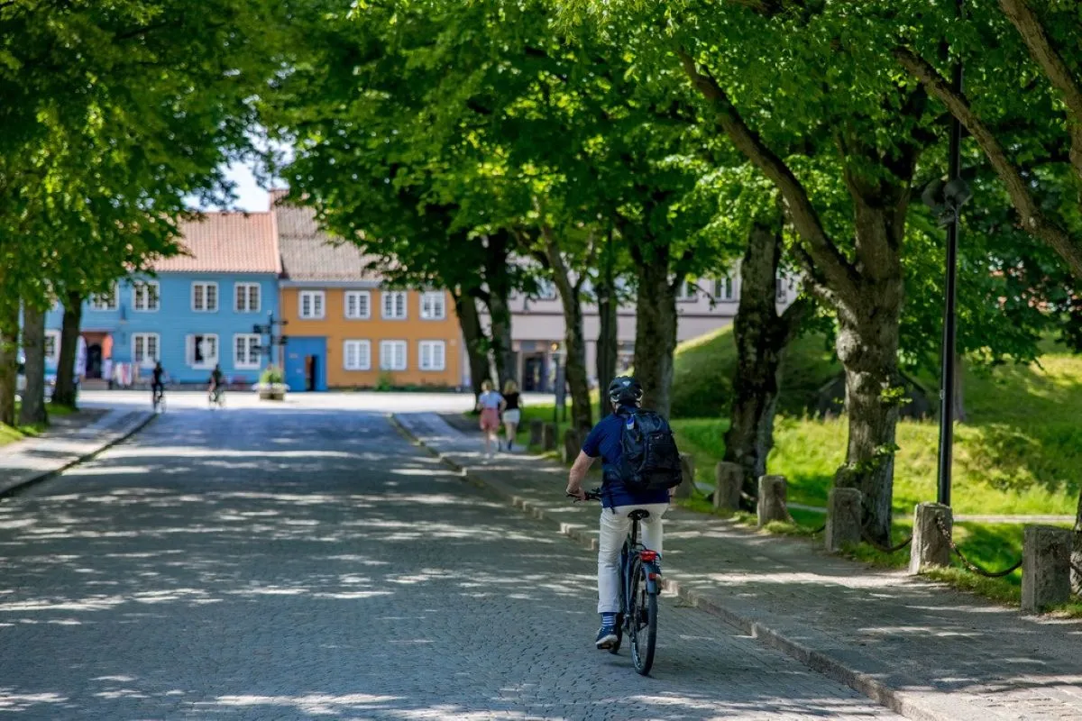 En syklist med hjelm og ryggsekk triller langs brosteinsgate under grønne trær, mot fargerike hus.
