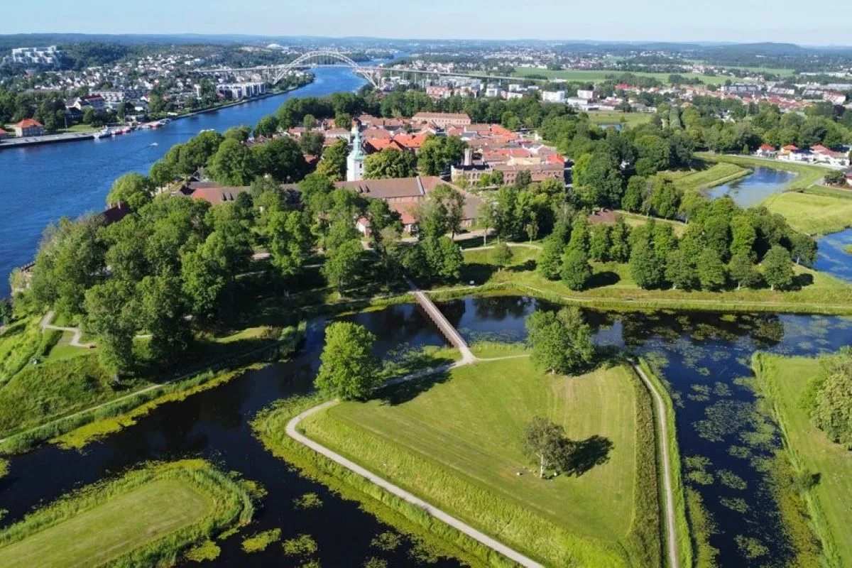 Luftfoto av elv og kanaler med parklandskap, broer og bygninger med røde tak, og by i bakgrunnen.