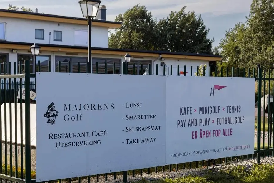 Stort informasjonsskilt på gjerde foran en bygning, med tekst om kafé, minigolf, tennis og matservering.