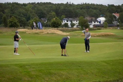 Tre golfspillere på en green; én putter mens de andre ser på, med trær og hus i bakgrunnen.