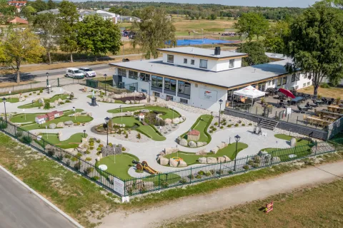 Luftfoto av inngjerdet minigolfbane med grønne baner og steiner, ved et klubbhus med uteservering.