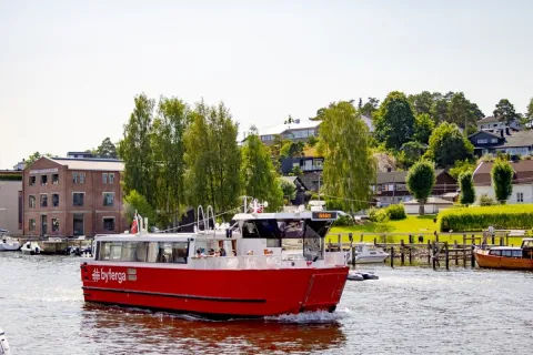 En rød passasjerbåt på vannet foran en grønn kyst med brygge, hus, trær og et murbygg i sollys.