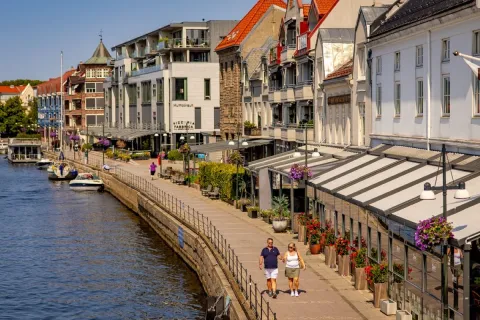 To personer går langs en kanalpromenade med kafeer, blomsterkasser og båter, foran bygårder i sollys.
